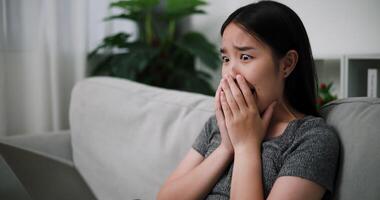 Handheld steady shot, Asian woman sitting on sofa looks shocked while watching something on laptop in living room at home. video