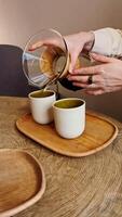 värma filtrera kaffe cascading från hantverkare glas och trä Häll i över uppstart, långsamt strömning in i ren vit muggar placerad på rustik trä- bricka, fångande noggrann bryggning artisteri video