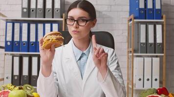 A healthcare professional examines a hamburger while surrounded by fresh fruits and vegetables, reflecting on dietary choices in a well-organized clinic environment. video