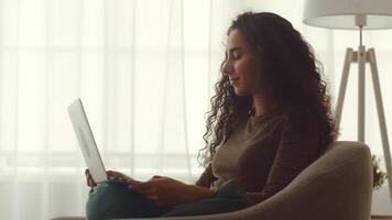 A young Middle Eastern woman enjoys her leisure time at home, comfortably seated while engaging with her laptop. Soft light filters through the curtains, creating a warm atmosphere. video