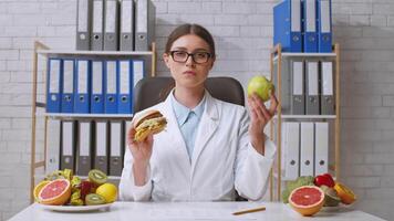 A dietitian evaluates a hamburger and an apple while seated at a desk in a modern clinic. Behind her, colorful fruits and organized files highlight the importance of nutrition. video