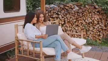 Two diverse girlfriends are seated together on a cozy bench in a camping area, looking at a laptop, enjoying each other's company on a sunny summer weekend with stacked logs in the background. video