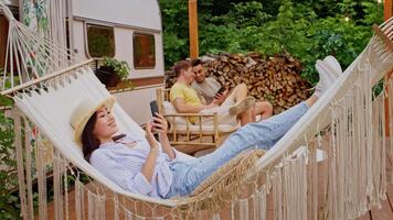 Group of diverse friends relax in a natural setting during summer, with one person on a hammock using a phone while others engage in light conversations, creating a joyful atmosphere. video