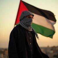 A man in a black jacket and a scarf stands in front of the palestinian flag photo