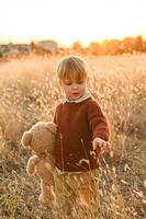 Dressed in old-fashioned clothes, a boy stands in tall grass at golden hour, gazing at the horizon with a large plush bear by his side. photo