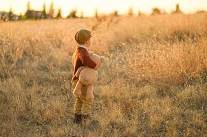 In the warm glow of sunset, a boy wearing vintage suspenders and a cap sits in a sunlit field, hugging his oversized teddy bear. photo