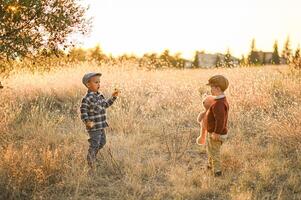 Two boys in vintage clothes play in a golden field at sunset, one holding a teddy bear, the other flying a small wooden airplane. photo