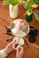 A woman sits in front of a mirror holding a blush compact, with her reflection visible as she prepares to apply it. photo