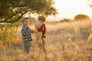 A peaceful rural sunset frames two boys in vintage attire, sharing a moment with a toy bear and a red apple in hand. photo
