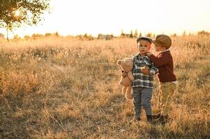Two boys dressed in vintage clothes stand in a golden field at sunset, one holding an apple and the other clutching a large teddy bear. photo