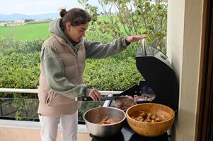 Next to a smoking barbecue, a woman in casual clothes stands on a modern terrace, with flavorful mushrooms and chicken ready to be grilled. photo