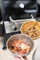 A pair of hands holds a platter with marinated chicken and mushrooms over a lit grill. photo