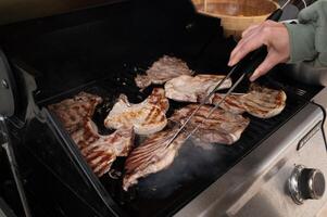 Outdoor grill filled with sizzling mixed meats, one piece being turned with tongs, surrounded by dancing flames and smoky atmosphere. photo