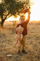 A little boy in a vintage cap stands in a golden field at sunset, holding an apple and a large teddy bear. photo