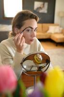 Woman shaping her brows using a brow pencil, seated at a table with makeup tools. photo