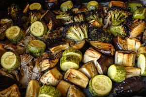 A tray of roasted vegetables with broccoli, eggplant, zucchini, and golden potatoes rests on the table, hot and ready to serve. photo