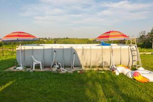 Above-ground pool in a grassy backyard with colorful umbrellas and inflatable toys, set in a rural landscape. Summer fun under the sun. photo
