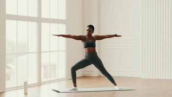 An African American woman performs a yoga pose in a well-lit room, showcasing her athleticism and commitment to a healthy lifestyle while enjoying her workout routine at home. video