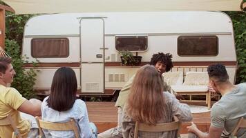 A diverse group of young adults engages in fun activities while resting together outdoors. They sit around a cozy area near a camper, enjoying the warmth of a summer weekend. video