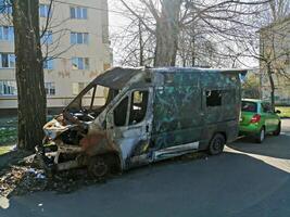 Kyiv, Ukraine - April 11, 2025 Heavily fire-damaged van with missing parts parked on residential street in front of apartment block. Urban decay and abandonment. Burned van near residential building photo