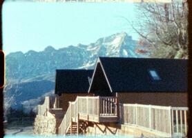 Wooden mountain chalet nestling before Pyrenees mountain range on clear spring morning, capturing nostalgic vintage 8mm film aesthetic with rustic wooden terrace and balcony video