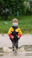 A small boy in a medical mask and rubber boots jumps through puddles on a spring day after a rain. Slow motion. video