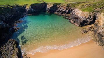 Secluded cove with clear turquoise water and sandy beach, framed by rocky cliffs and green grass. photo