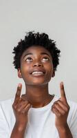 Smiling young black woman in white t-shirt looking and pointing up with both hands. Confident and friendly expression. Suitable for advertising and promotion. photo
