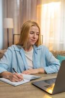 Young woman studying at home laptop lesson focus, notes in notebook, attentive learning moment photo