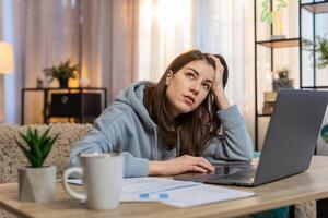Tired young woman freelancer using laptop computer at table with documents in living room at home photo