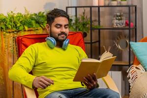 Indian man reading interesting book turning pages smiling enjoying literature taking a rest on chair photo