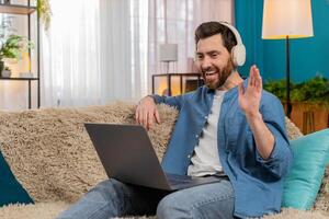 Young man in wireless headphones waving at webcam, laughing while talking on call using laptop photo