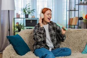Woman sitting on sofa wearing headphones singing with comb as microphone having fun with music joy photo