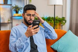 Shocked sad Indian young man using smartphone reading negative message feels annoyed app crash photo