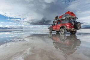A rugged four-wheel drive vehicle with a rooftop tent stands on the expansive salt flats, reflecting the clouds above. The vast landscape showcases beautiful blues and whites during a cloudy day. photo
