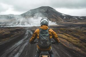 A motorcyclist wearing a helmet and a backpack navigates a muddy path across a volcanic landscape in Iceland. Steam rises from the ground, hinting at geothermal activity nearby, under an overcast sky. photo