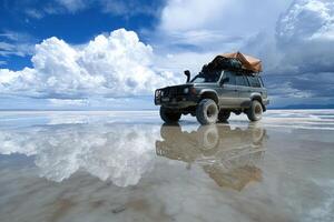 A rugged off-road vehicle is parked on a vast salt flat under a sky filled with dramatic clouds. The ground reflects the vehicle and clouds, creating a stunning visual contrast. photo