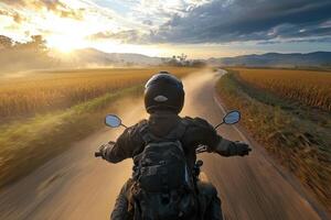 A motorcyclist navigates a winding road as the sun sets. Dust rises from the tires, creating a hazy atmosphere. Lush fields border the path, enhancing the tranquil beauty of the scene. photo