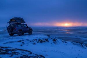 A rugged vehicle is parked on a rocky outcrop, overlooking a snowy landscape at sunset. The vibrant colors of the sky blend beautifully as night approaches in this remote winter setting. photo