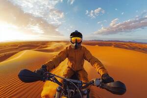 A rider clad in an orange jumpsuit navigates a motorcycle over vast, undulating sand dunes during sunset. The sky is filled with clouds and warm hues, showcasing a serene desert atmosphere. photo
