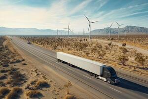 A large truck navigates a highway through a dry desert landscape. Wind turbines line the horizon, highlighting renewable energy. The sun casts a warm glow over the empty road and distant mountains. photo
