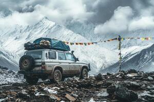 A rugged vehicle stands on rocky terrain in a snowy mountain landscape, surrounded by dramatic peaks and clouds. Colorful prayer flags flutter in the breeze, adding vibrancy to the serene scenery. photo
