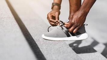 Black sportsman tying shoelaces before running, getting ready for jogging outdoors, putting leg at steps, cropped image with copy space, side view photo