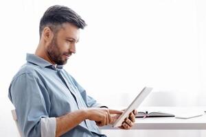 Handsome Freelancer Man Using Digital Tablet While Having Break At Work, Sitting At Desk In Home Office, Browsing Internet Or Social Networks, Checking App, Working Online, Side View With Copy Space photo