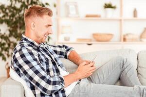 Enjoying Day Off. Side view portrait of casual guy using his smartphone, resting on the couch at home, blurred background, free space. Smiling man chatting online, browsing social media, texting sms photo