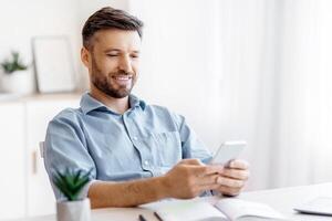 Handsome Young Businessman Using Smartphone At Workplace In Office, Checking New Application, Browsing Internet, Messaging With Friends, Playing Online Games While Having Break At Work, Free Space photo