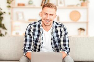 Portrait Of Handsome Confident Bearded Guy In Shirt Sitting On The Sofa At Home, Looking At Camera And Smiling. Happy Man Working On Computer, Copy Space, Blurred Background, Indoors photo