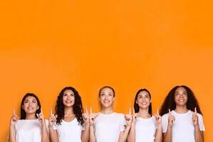 Look Upward. Five Multiethnic Women Pointing Fingers Up Posing Standing In Studio Over Yellow Background. Look There Advertisement, Females Showing Copy Space Above Head photo