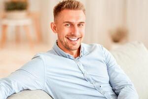 Portrait Of Handsome Millennial Bearded Man In Shirt Sitting And Leaning On Comfortable Couch In Living Room, Posing For Photo And Looking At Camera, Copy Space, Blurred Background, Indoors