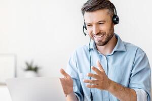 Conference. Friendly man office employee in headset having web call on laptop at workplace, talking and gesturing at camera, copy space photo